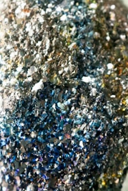 A close-up of a surface covered in sparkling, crystalline structures with various hues of blue, silver, and hints of gold, giving a metallic and textured appearance.