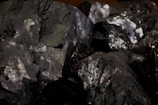 A collection of dark, metallic minerals with shiny surfaces and various angular fragments. The texture appears rough with a mix of reflective and matte areas.