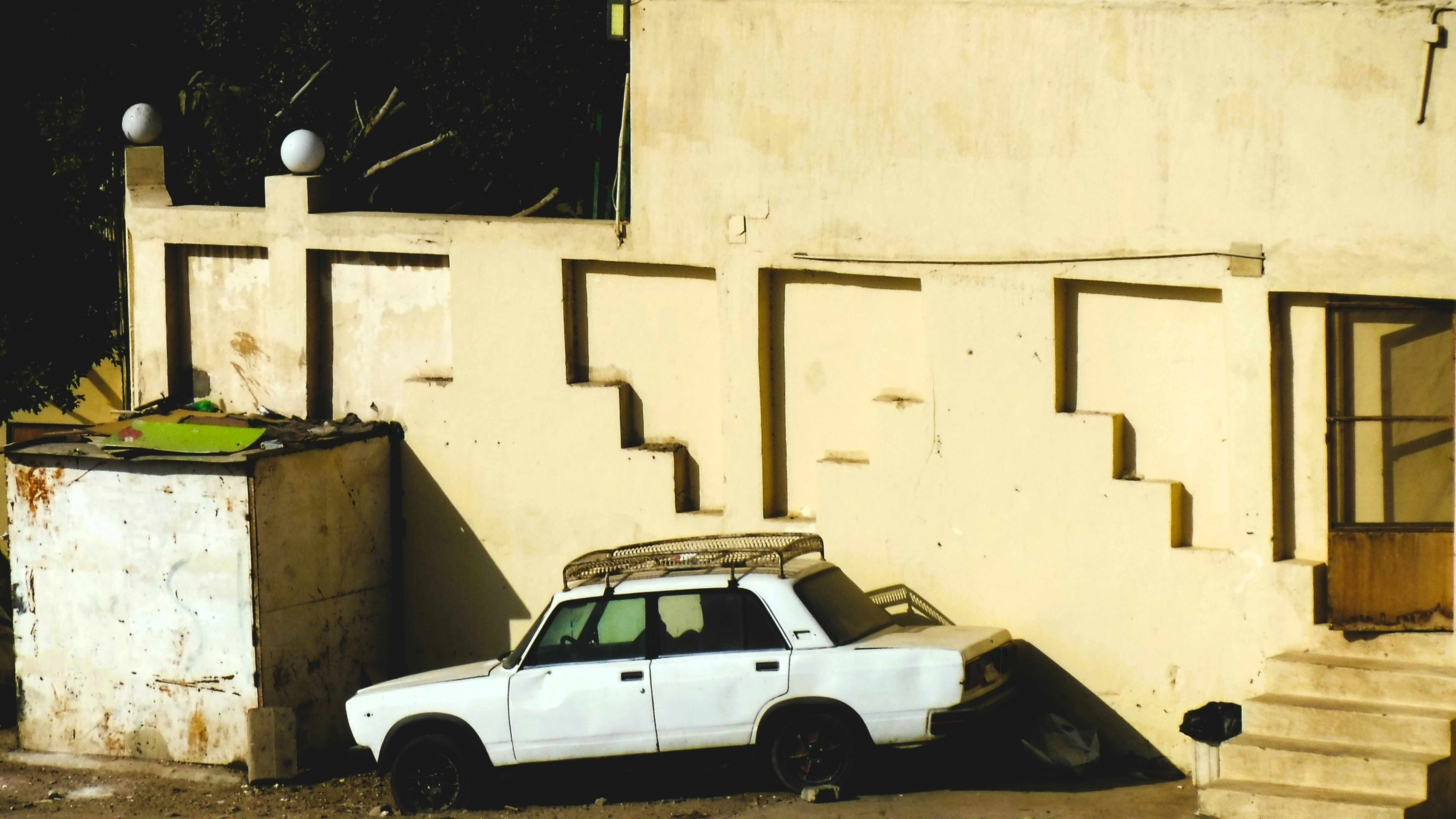 White sedan pressed against a sunlit stair-step wall beside a weathered beige building. The scene emphasizes geometric shadows and urban texture.