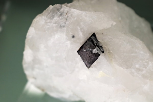 A large crystal with a white, translucent appearance. It has a small, dark, metallic-looking mineral embedded in its surface, displaying a cubic shape. The texture of the crystal appears rough with natural grooves and imperfections.