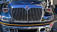 The front view of a large, polished truck with a shiny chrome grille and headlight details, reflecting its urban surroundings. People are visible in the background on the sidewalk.