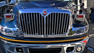 The front view of a large, polished truck with a shiny chrome grille and headlight details, reflecting its urban surroundings. People are visible in the background on the sidewalk.