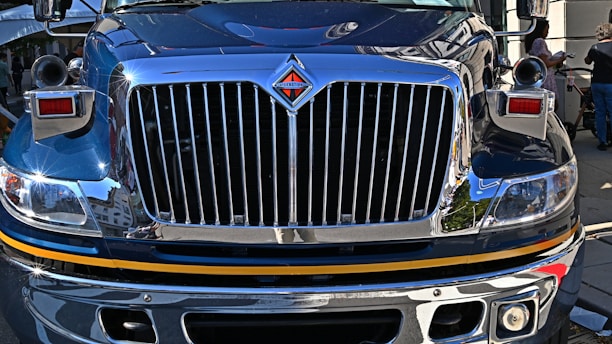 The front view of a large, polished truck with a shiny chrome grille and headlight details, reflecting its urban surroundings. People are visible in the background on the sidewalk.