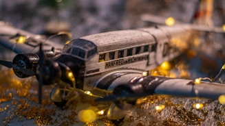 A model airplane is decorated with small, warm white string lights. The plane has a vintage design with detailed markings. It is set on a surface with gold and white accents, creating a festive and nostalgic atmosphere.