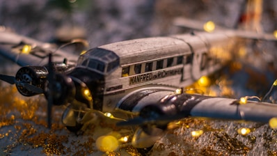 A model airplane is decorated with small, warm white string lights. The plane has a vintage design with detailed markings. It is set on a surface with gold and white accents, creating a festive and nostalgic atmosphere.
