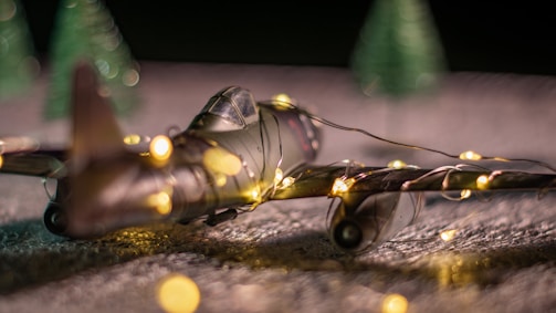 A model airplane wrapped in string lights rests on a textured surface, with small, blurry, artificial green trees positioned in the background. The scene is softly lit, creating a cozy, festive ambiance.