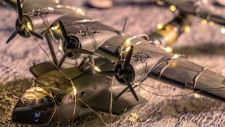 A scale model of a vintage aircraft adorned with string lights, positioned on a textured surface. The aircraft features four propellers and is bathed in warm, ambient lighting.