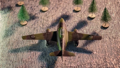 A model of a vintage military aircraft is positioned on a textured surface. The aircraft is surrounded by five miniature pine trees on small wooden bases. The plane has a camouflage pattern with insignia on the wings.