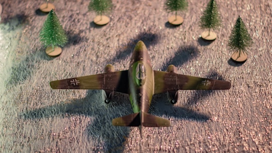 A model of a vintage military aircraft is positioned on a textured surface. The aircraft is surrounded by five miniature pine trees on small wooden bases. The plane has a camouflage pattern with insignia on the wings.
