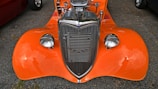 An orange vintage car with a polished chrome grille and high-performance engine visible under the open hood. The car is parked on an asphalt surface with reflections visible on the shiny surface of the body and headlights.