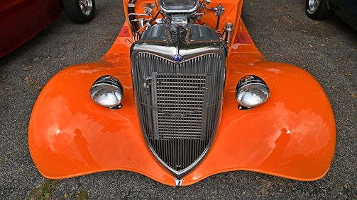 An orange vintage car with a polished chrome grille and high-performance engine visible under the open hood. The car is parked on an asphalt surface with reflections visible on the shiny surface of the body and headlights.