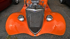 An orange vintage car with a polished chrome grille and high-performance engine visible under the open hood. The car is parked on an asphalt surface with reflections visible on the shiny surface of the body and headlights.