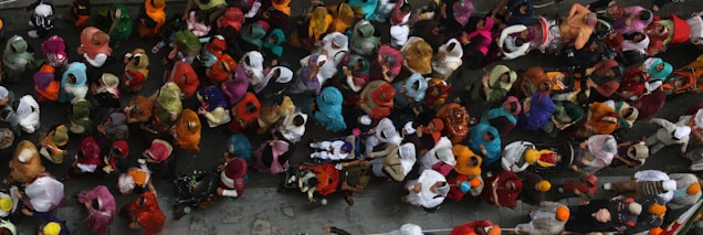 A vibrant crowd seen from above, with individuals wearing colorful traditional clothing such as saris and turbans. The scene is dense with people of various ages, suggesting a festive or ceremonial gathering. The clothing ranges in hues from bright orange and red to deep blue and green, creating a lively and dynamic mosaic.