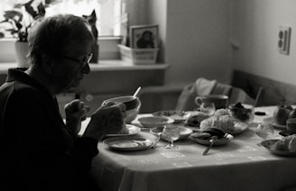 An elderly person sits at a dining table, holding a cup. The table is set with various dishes, pastries, and cups, indicating a meal. The environment appears cozy with natural light coming in through a window, and a houseplant is visible in the background.