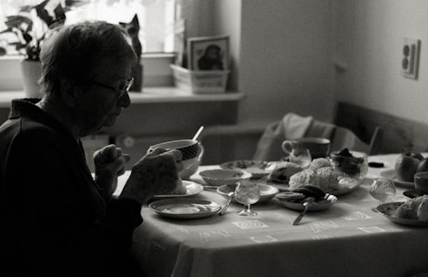 An elderly person sits at a dining table, holding a cup. The table is set with various dishes, pastries, and cups, indicating a meal. The environment appears cozy with natural light coming in through a window, and a houseplant is visible in the background.