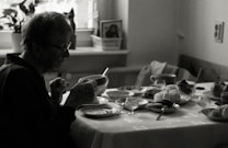 An elderly person sits at a dining table, holding a cup. The table is set with various dishes, pastries, and cups, indicating a meal. The environment appears cozy with natural light coming in through a window, and a houseplant is visible in the background.