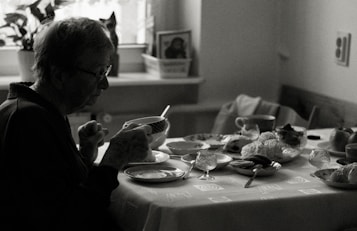 An elderly person sits at a dining table, holding a cup. The table is set with various dishes, pastries, and cups, indicating a meal. The environment appears cozy with natural light coming in through a window, and a houseplant is visible in the background.