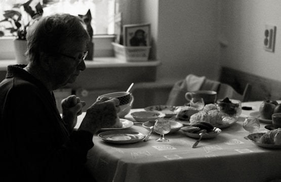 An elderly person sits at a dining table, holding a cup. The table is set with various dishes, pastries, and cups, indicating a meal. The environment appears cozy with natural light coming in through a window, and a houseplant is visible in the background.