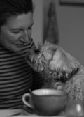 A person with a gentle smile leans towards a small, fluffy dog, creating an intimate and warm moment. They appear to be sharing a close connection. In the foreground, there is a cup, possibly suggesting a cozy setting.