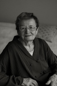 An elderly person with glasses, wearing a dark patterned sweater, sits calmly with a slight smile. The background is softly blurred, adding to the focus on the person's gentle expression and wise demeanor.