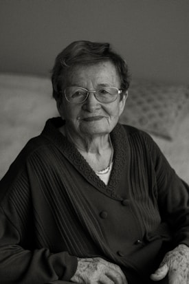 An elderly person with glasses, wearing a dark patterned sweater, sits calmly with a slight smile. The background is softly blurred, adding to the focus on the person's gentle expression and wise demeanor.