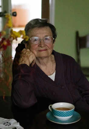 A warm scene of an elderly woman smiling gently while a friendly caregiver assists her with a cup of tea at home.