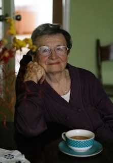 An elderly woman with glasses is sitting at a table, resting her head on her hand. She is wearing a dark sweater and has a gentle expression. In front of her is a turquoise cup filled with coffee or tea. The background is a soft green, with a blurred glimpse of furniture and yellow flowers, adding a warm, homely atmosphere.