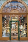 A visually appealing storefront showcasing a variety of decorative items. The window display features twinkling lights, paper stars, and ornately arranged plants and ornaments. A festive ambiance is accentuated by the garland framing the entrance. Above the door, a large window with an arched design provides a glimpse of the glass ceiling and structure beyond, adding depth and interest.