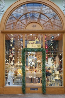 A visually appealing storefront showcasing a variety of decorative items. The window display features twinkling lights, paper stars, and ornately arranged plants and ornaments. A festive ambiance is accentuated by the garland framing the entrance. Above the door, a large window with an arched design provides a glimpse of the glass ceiling and structure beyond, adding depth and interest.
