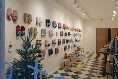 A well-organized display of colorful slippers hanging on a white wall in a cozy indoor setting. A small evergreen tree sits in the corner, adding a festive touch. The floor features a black and white checkered pattern, and benches display more slippers and related items. Warm lighting from ceiling lights enhances the inviting ambiance.