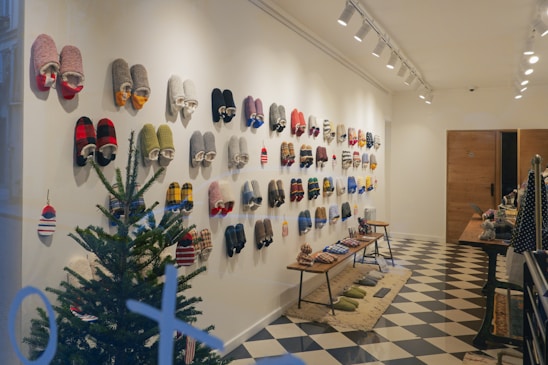 A well-organized display of colorful slippers hanging on a white wall in a cozy indoor setting. A small evergreen tree sits in the corner, adding a festive touch. The floor features a black and white checkered pattern, and benches display more slippers and related items. Warm lighting from ceiling lights enhances the inviting ambiance.