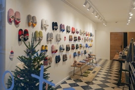 A well-organized display of colorful slippers hanging on a white wall in a cozy indoor setting. A small evergreen tree sits in the corner, adding a festive touch. The floor features a black and white checkered pattern, and benches display more slippers and related items. Warm lighting from ceiling lights enhances the inviting ambiance.