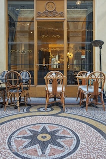 A quaint outdoor Parisian cafe with vintage wooden chairs and tables arranged on a stone mosaic floor. The mosaic features a circular star pattern in shades of black, yellow, and terracotta. The cafe, named 'Le Valentin', is fronted by a large glass door with intricate designs above. Reflections of the interior lights add a warm glow to the scene.