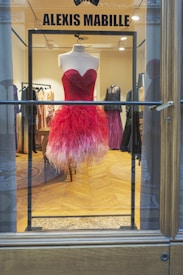 A vibrant red and pink feathered dress displayed on a mannequin inside a high-end fashion boutique. The dress has a heart-shaped neckline and a flamboyant feathered skirt. The background showcases other elegant gowns in various shades, creating a luxurious and sophisticated ambiance.