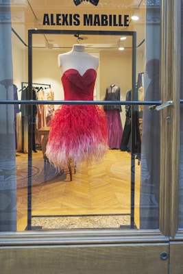 A vibrant red and pink feathered dress displayed on a mannequin inside a high-end fashion boutique. The dress has a heart-shaped neckline and a flamboyant feathered skirt. The background showcases other elegant gowns in various shades, creating a luxurious and sophisticated ambiance.