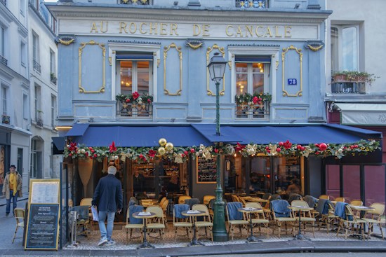 A quaint street-side caf&eacute; decorated for the holiday season. The exterior of the building is adorned with festive red, gold, and green decorations. There are numerous empty chairs and tables set up outside under a navy blue awning. A few people are visible, one walking towards the entrance and another standing at the side. The caf&eacute; has a classic European architecture with ornate moldings and a sign reading 'Au Rocher de Cancale' across the top.