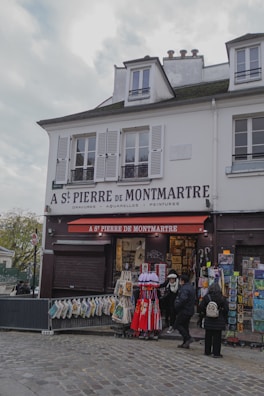 A quaint Parisian storefront named A St Pierre de Montmartre, offering art supplies and souvenirs. The facade displays various artworks and bags for sale. A few people are browsing the items outside under a red awning. The building is white with shuttered windows and has a charming, historical architecture.