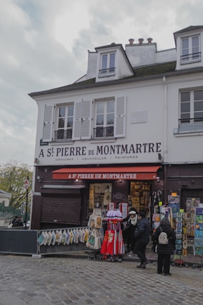 A quaint Parisian storefront named A St Pierre de Montmartre, offering art supplies and souvenirs. The facade displays various artworks and bags for sale. A few people are browsing the items outside under a red awning. The building is white with shuttered windows and has a charming, historical architecture.