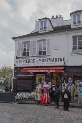 A quaint Parisian storefront named A St Pierre de Montmartre, offering art supplies and souvenirs. The facade displays various artworks and bags for sale. A few people are browsing the items outside under a red awning. The building is white with shuttered windows and has a charming, historical architecture.