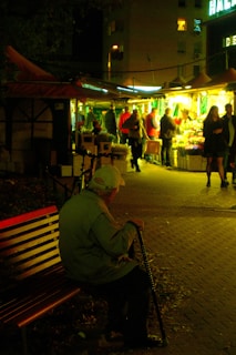 An elderly person is sitting alone on a bench in a dimly-lit area, holding a walking cane. In the background, there is a lively market stall illuminated with bright lights where people are gathered, shopping for flowers and other goods. The atmosphere contrasts between the solitude of the person on the bench and the bustling market scene.