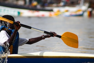 A person is paddling a canoe or kayak on a body of water. The individual is wearing a life vest and gripping a yellow paddle. The water around the paddle displays splashes, indicating movement. There are blurred boats and colorful reflections in the backdrop.