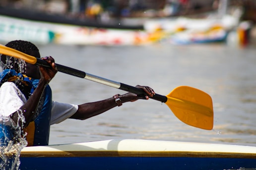 A person is paddling a canoe or kayak on a body of water. The individual is wearing a life vest and gripping a yellow paddle. The water around the paddle displays splashes, indicating movement. There are blurred boats and colorful reflections in the backdrop.