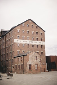 A large, old brick warehouse building with multiple small square windows and a smaller attached structure. The building has a sign that reads 'Vinings Warehouse'. The area is deserted, with a couple of empty benches in the foreground, and the sky is overcast.