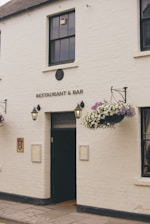 A white brick building with the sign 'RESTAURANT & BAR' above a black door. Hanging baskets with flowers are mounted on either side of the entrance. Two lantern-style lights are affixed to the wall near the door. The building has a traditional appearance with quaint, decorative touches.