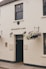A white brick building with the sign 'RESTAURANT & BAR' above a black door. Hanging baskets with flowers are mounted on either side of the entrance. Two lantern-style lights are affixed to the wall near the door. The building has a traditional appearance with quaint, decorative touches.