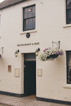 A white brick building with the sign 'RESTAURANT & BAR' above a black door. Hanging baskets with flowers are mounted on either side of the entrance. Two lantern-style lights are affixed to the wall near the door. The building has a traditional appearance with quaint, decorative touches.