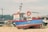 A small, colorful fishing boat is positioned on a pebble-strewn beach. The blue boat features a bright red trim and has the label 'R4' on its side. A lifebuoy is mounted on the side railing, and a small cabin is visible with windows. Behind the boat, various indistinct objects and pieces of equipment are scattered, suggesting a working area or storage yard. In the background, there are trees and buildings, indicating a coastal or dockside environment under a clear sky.