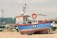 A small, colorful fishing boat is positioned on a pebble-strewn beach. The blue boat features a bright red trim and has the label 'R4' on its side. A lifebuoy is mounted on the side railing, and a small cabin is visible with windows. Behind the boat, various indistinct objects and pieces of equipment are scattered, suggesting a working area or storage yard. In the background, there are trees and buildings, indicating a coastal or dockside environment under a clear sky.