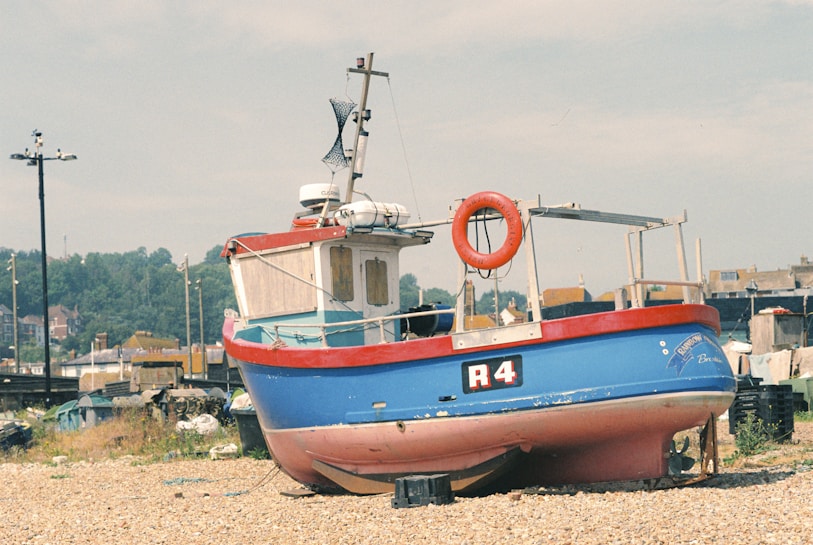 A small, colorful fishing boat is positioned on a pebble-strewn beach. The blue boat features a bright red trim and has the label 'R4' on its side. A lifebuoy is mounted on the side railing, and a small cabin is visible with windows. Behind the boat, various indistinct objects and pieces of equipment are scattered, suggesting a working area or storage yard. In the background, there are trees and buildings, indicating a coastal or dockside environment under a clear sky.