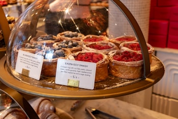 A display of pastries under a glass dome, featuring raspberry and blueberry Danish pastries with flaky pastry, almond cream, vanilla ganache, and a sugar dusting. Price tags are visible, showing each pastry priced at $5.9.